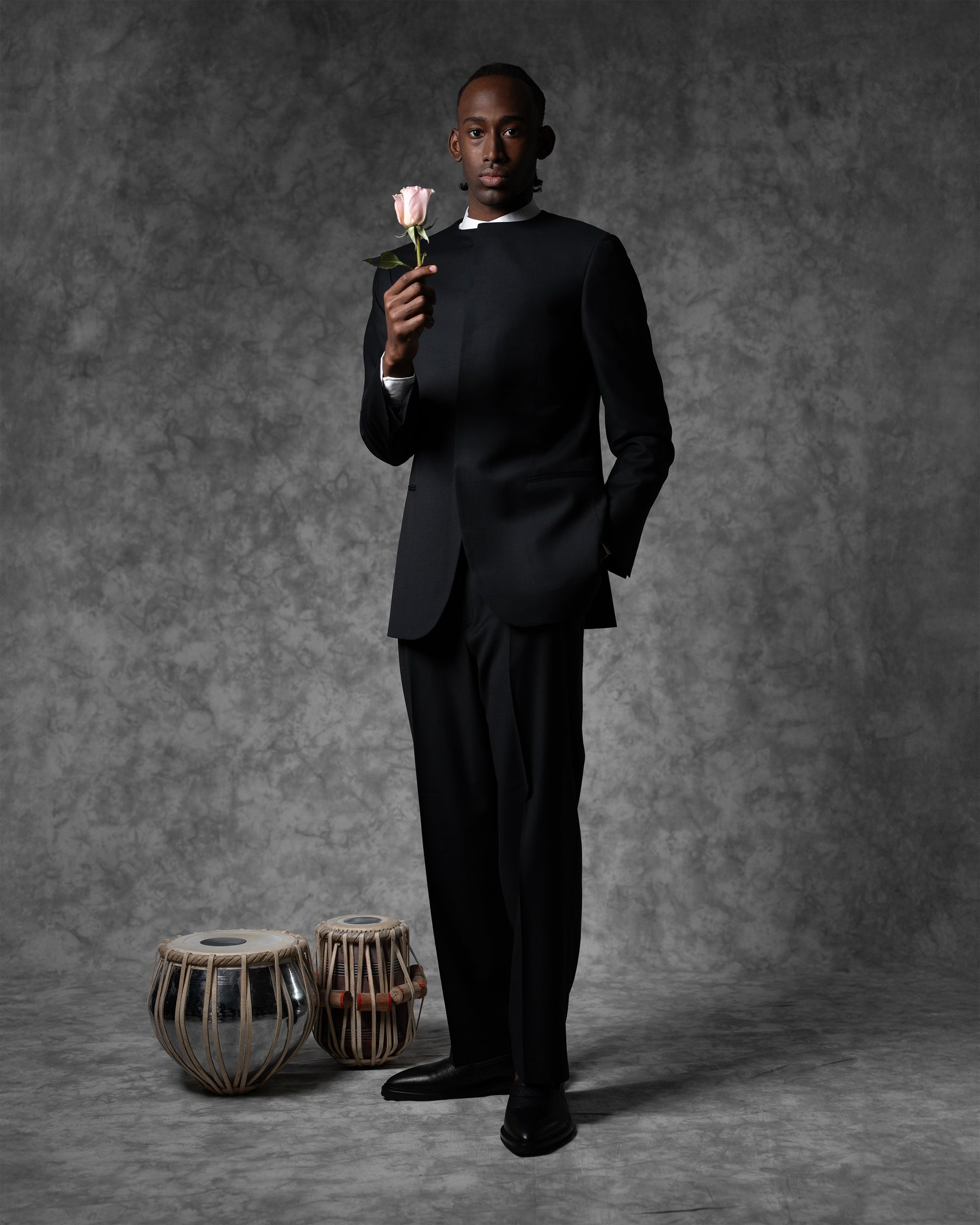 Man in a black suit holding a rose against a gray background with drums.